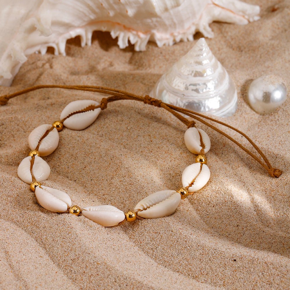 Bracelet de cheville marron en corde avec coquillages blancs et perles dorées sur sable fin.