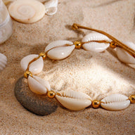 Bracelet de cheville en corde marron avec coquillages blancs et perles dorées posé sur du sable.
