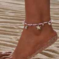 Bracelet de cheville rose avec perles, coquillages et étoile de mer en résine sur peau bronzée plage sable fin.
