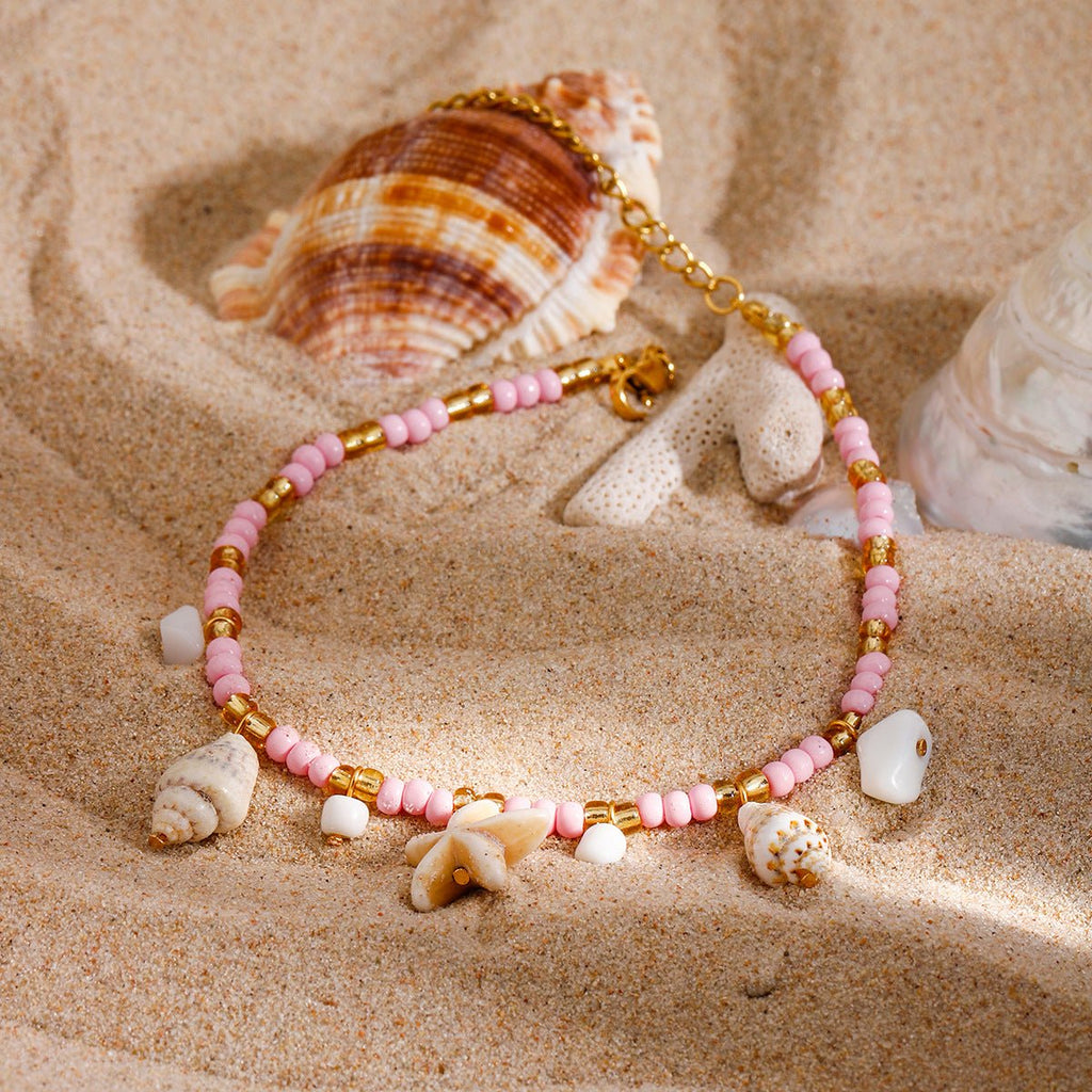 Bracelet de cheville rose avec perles, coquillages et étoile de mer en résine sur sable.