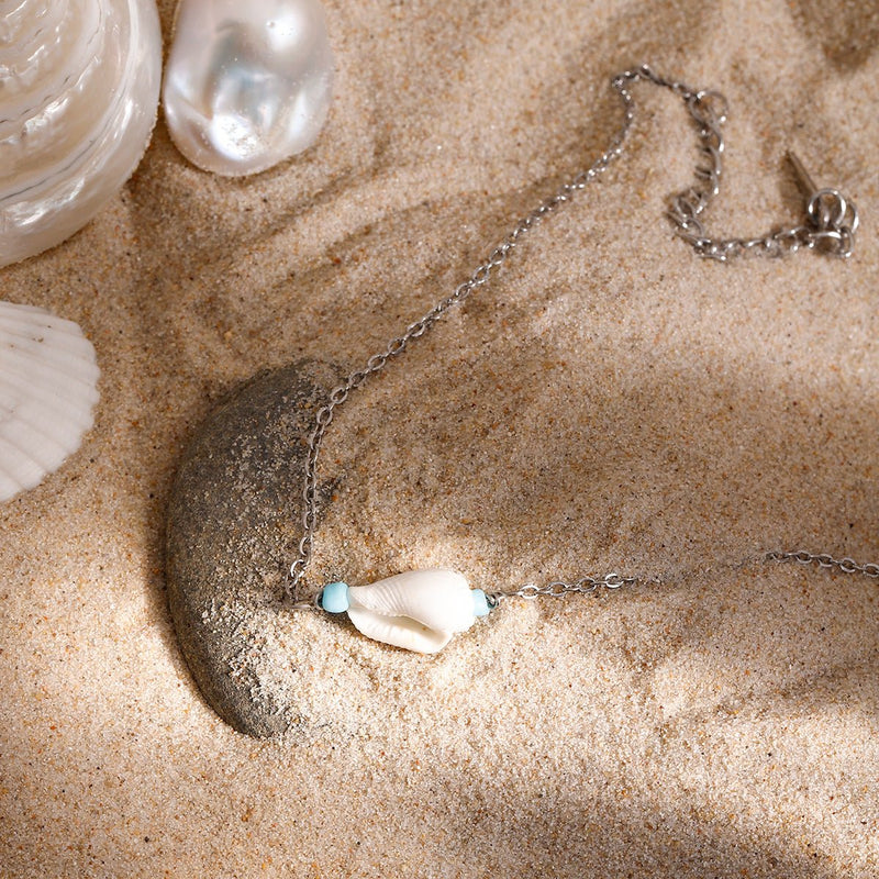 Bracelet de cheville double en acier inoxydable argenté avec coquillage et perle turquoise sur sable.