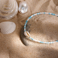Bracelet de cheville double en acier inoxydable avec perles bleues, blanches et coquillage naturel sur sable fin.