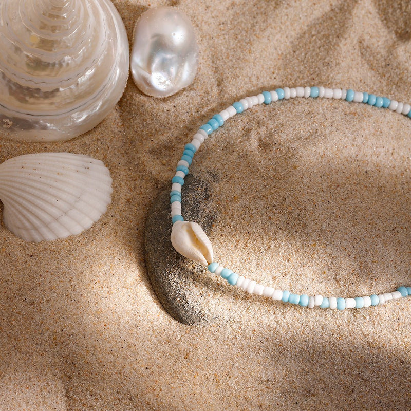 Bracelet de cheville double en acier inoxydable avec perles bleues, blanches et coquillage naturel sur sable fin.