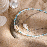 Bracelet de cheville double en acier inoxydable avec coquillages blancs et perles bleues et blanches sur sable beige.