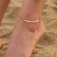 Bracelet de cheville double rang en perles d'eau douce blanches et acier inoxydable doré porté sur pied féminin sur sable.