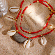 Bracelet de cheville rouge double tour en résine avec coquillage blanc et perles dorées sur sable fin.