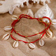 Bracelet de cheville double tour en fil rouge avec perles dorées et coquillages blancs posé sur sable.