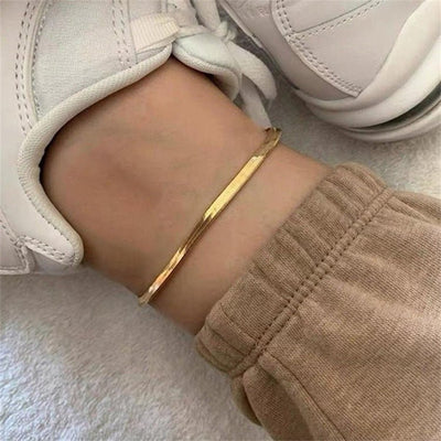 Bracelet de cheville en acier inoxydable doré, maille serpent lisse, porté avec baskets blanches et pantalon beige.