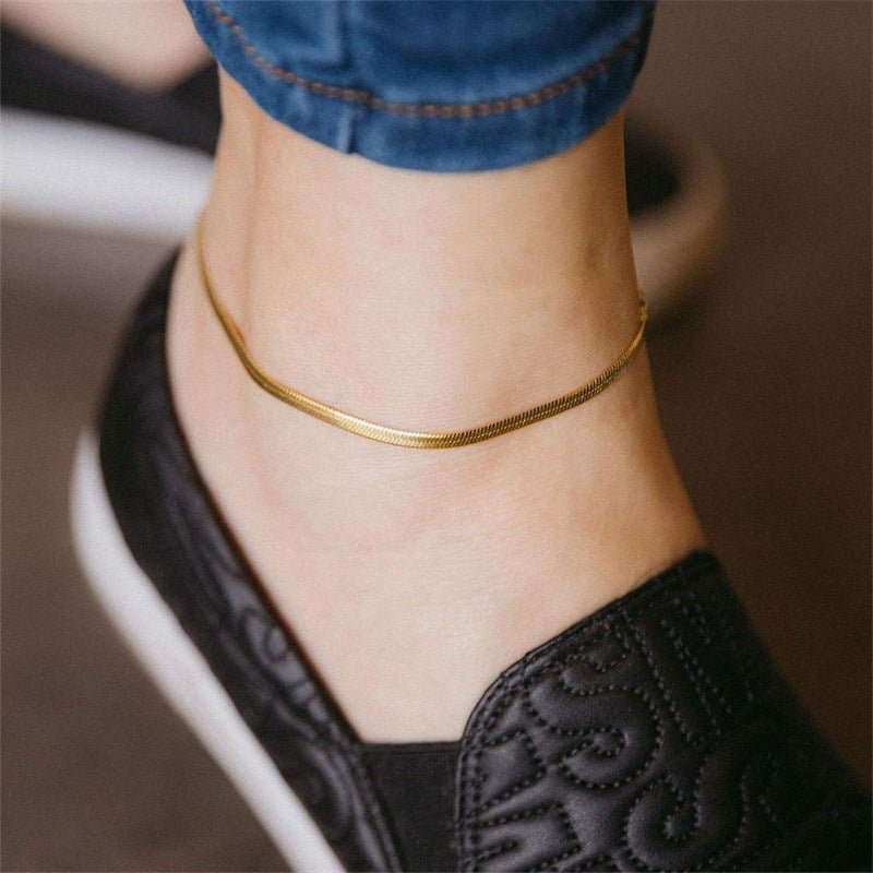 Bracelet de cheville en acier inoxydable doré, maille serpent lisse, porté sur pied féminin avec chaussure noire brodée.