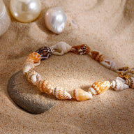 Bracelet de cheville en acier orné de coquillages naturels multicolores posé sur sable et pierre grise.
