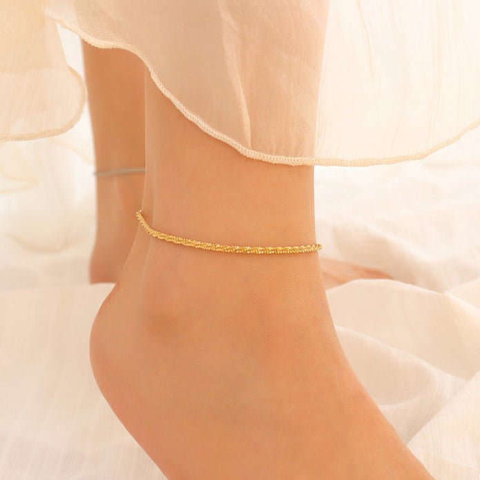 Bracelet de cheville en titane or à maille scintillante, modèle Lamina, porté sur peau pâle.