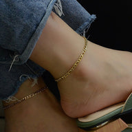 Bracelet de cheville en titane doré, maille figaro fine, porté avec jean déchiré et sandale beige.