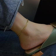 Bracelet de cheville en titane or, maille figaro fine, porté sur cheville féminine avec pantalon en jean et sandale verte.