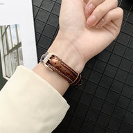 Bracelet Apple Watch en cuir marron foncé, couture beige, boucle argentée, porté au poignet féminin.