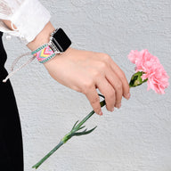 Bracelet Apple Watch en nylon rose avec cristaux multicolores, porté au poignet féminin, tenant une fleur rose.