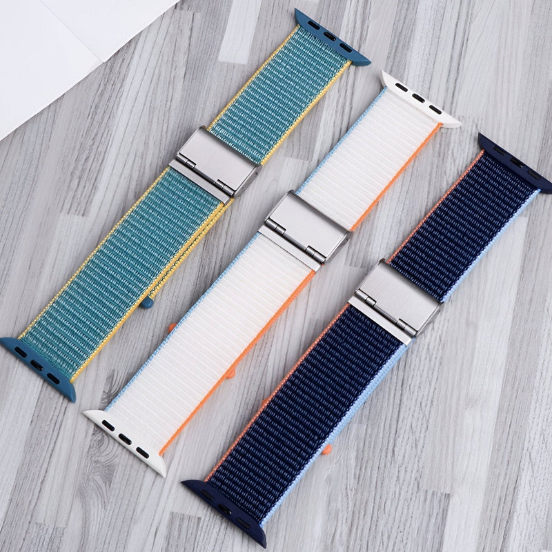 Trois bracelets Apple Watch en nylon, couleurs bleu-vert, blanc-orange, et bleu marine, boucles argentées.