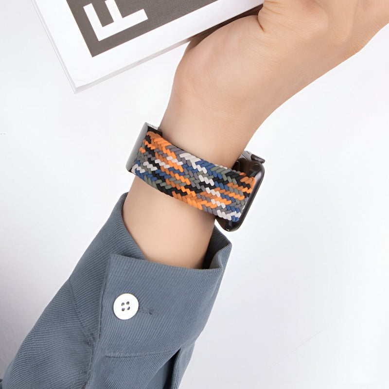 Bracelet Apple Watch en nylon tressé orange, noir et beige, fermé sur poignet avec chemise grise.
