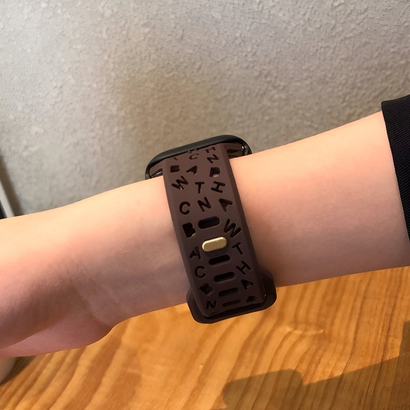 Bracelet Apple Watch silicone marron foncé avec motifs lettres noires, porté au poignet.
