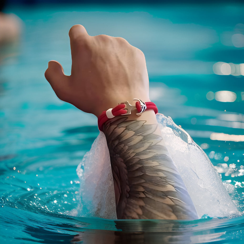 Bracelet double cordon rouge avec motif ancre argentée porté au poignet homme tatoué près de l'eau.