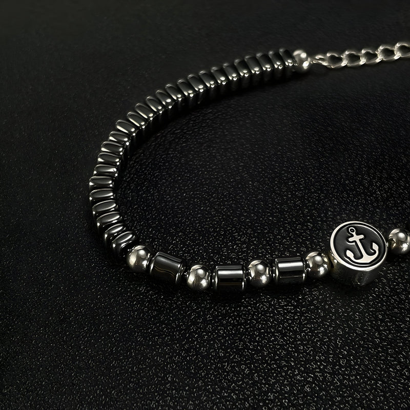 Bracelet homme en acier inoxydable et hématites noires avec charm ancre argenté sur fond noir.
