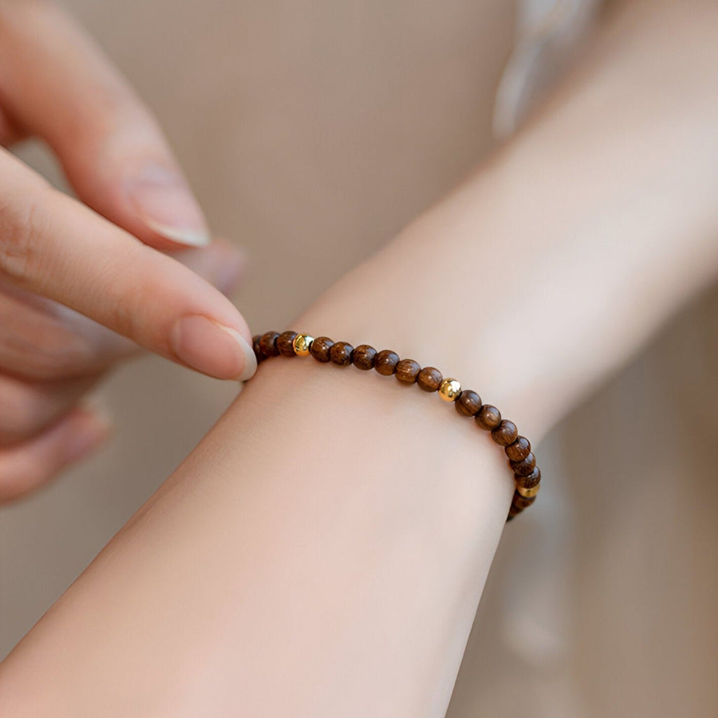 Bracelet fin en perles rondes marron en bois de santal avec une perle dorée sur poignet féminin.