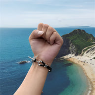 Bracelet en cuir noir avec avion en acier inoxydable porté au poignet masculin, bord de mer en arrière-plan.