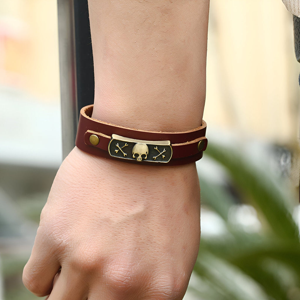 Bracelet en cuir marron double tour avec motif tête de mort et os croisés en métal doré sur plaque noire.