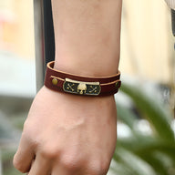 Bracelet en cuir marron double tour avec motif tête de mort et os croisés en métal doré sur plaque noire.