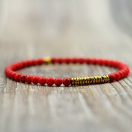 Bracelet fin pour homme en petites perles rouges et détails cylindriques dorés sur fond bois clair.