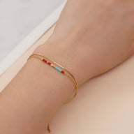 Bracelet double-rangs en perles rouges, bleues et blanches avec chaîne fine dorée sur poignet féminin.