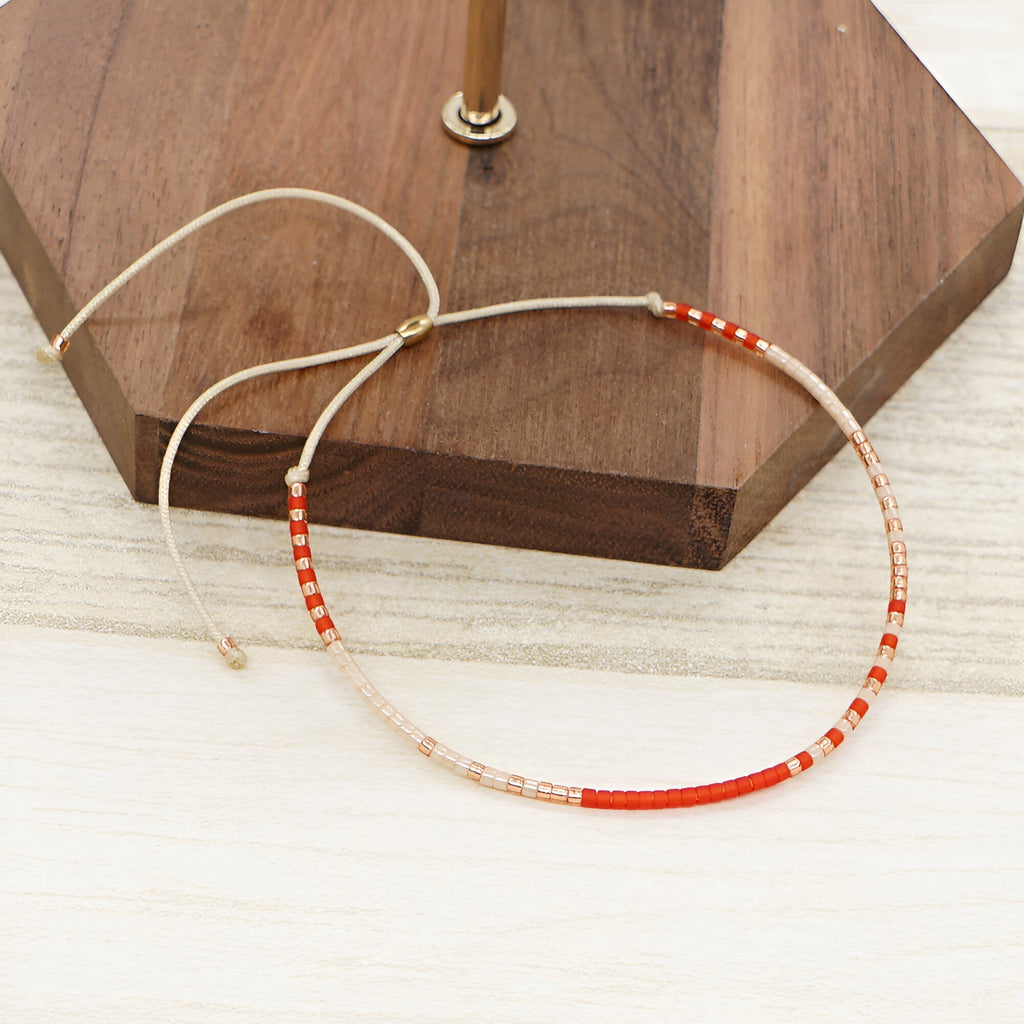 Bracelet fin en perles rouges et transparentes sur fil beige ajustable sur fond bois clair.