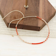 Bracelet fin en perles rouges et transparentes sur fil beige ajustable sur fond bois clair.