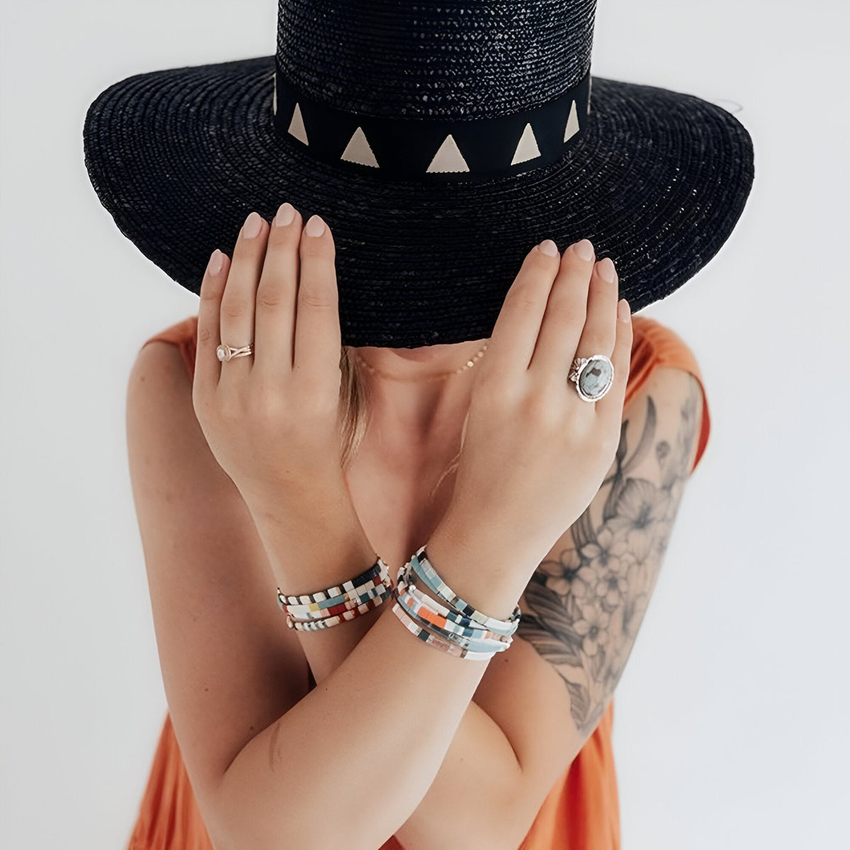 Bracelet multicolore en perles fines du Japon porté au poignet féminin avec bagues argentées et chapeau noir large à.