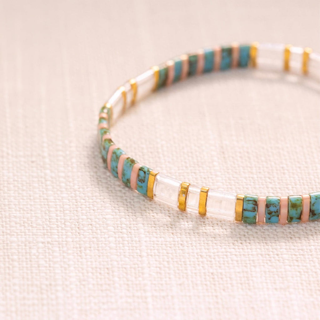 Bracelet fin en perles japonaises bleu turquoise, transparentes et blanches avec détails dorés.