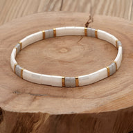 Bracelet femme perles blanches tubulaires avec fines bandes dorées sur bois naturel.