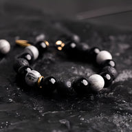Bracelet homme en pierres noires volcaniques et blanches avec détails dorés sur fond noir texturé.
