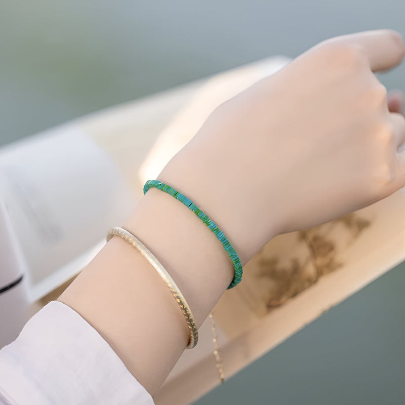 Bracelet fin en pierres turquoises vertes avec un autre bracelet doré tressé, porté au poignet féminin.