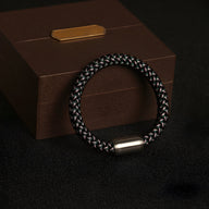 Bracelet en polyester tressé noir et gris avec fermoir cylindrique argenté sur fond noir et coffret marron.