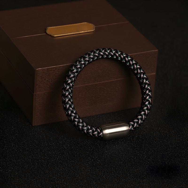 Bracelet en polyester tressé noir et gris avec fermoir cylindrique argenté sur fond noir et coffret marron.