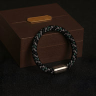 Bracelet unisexe en polyester noir et gris tressé avec fermoir métallique argenté sur boîte en bois foncé.