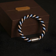 Bracelet tressé polyester noir, bleu et beige avec fermoir métallique argenté posé sur boîte marron.