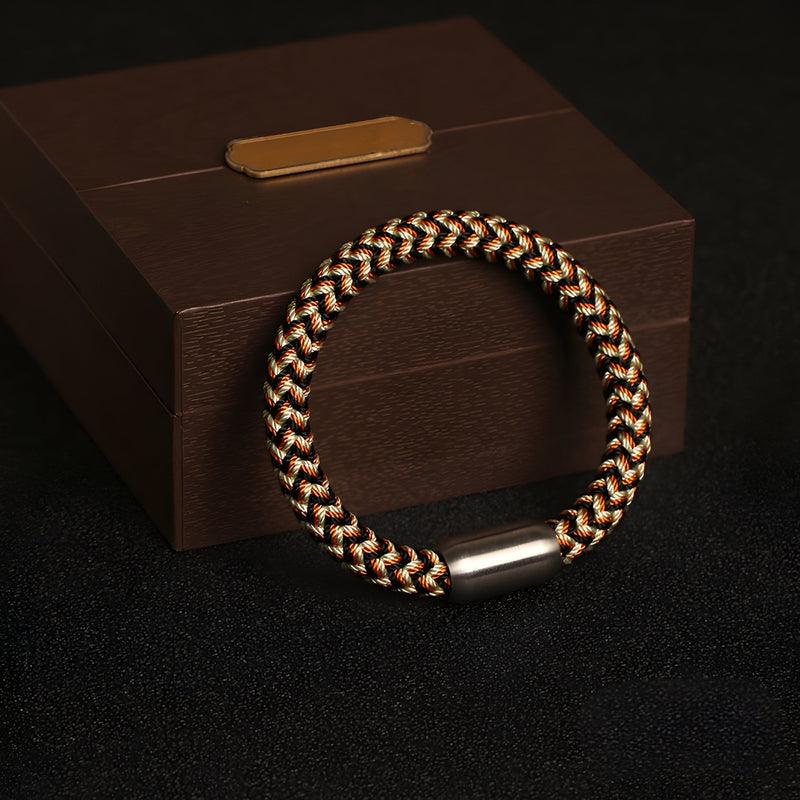 Bracelet tressé en polyester noir, beige et marron avec fermoir argenté sur boîte marron élégante.