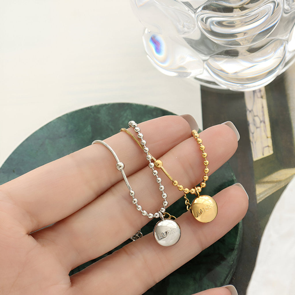 Bracelet en titane plaqué or 18 carats et argent, médaillon rond gravé "Lucky", chaîne à billes fines.