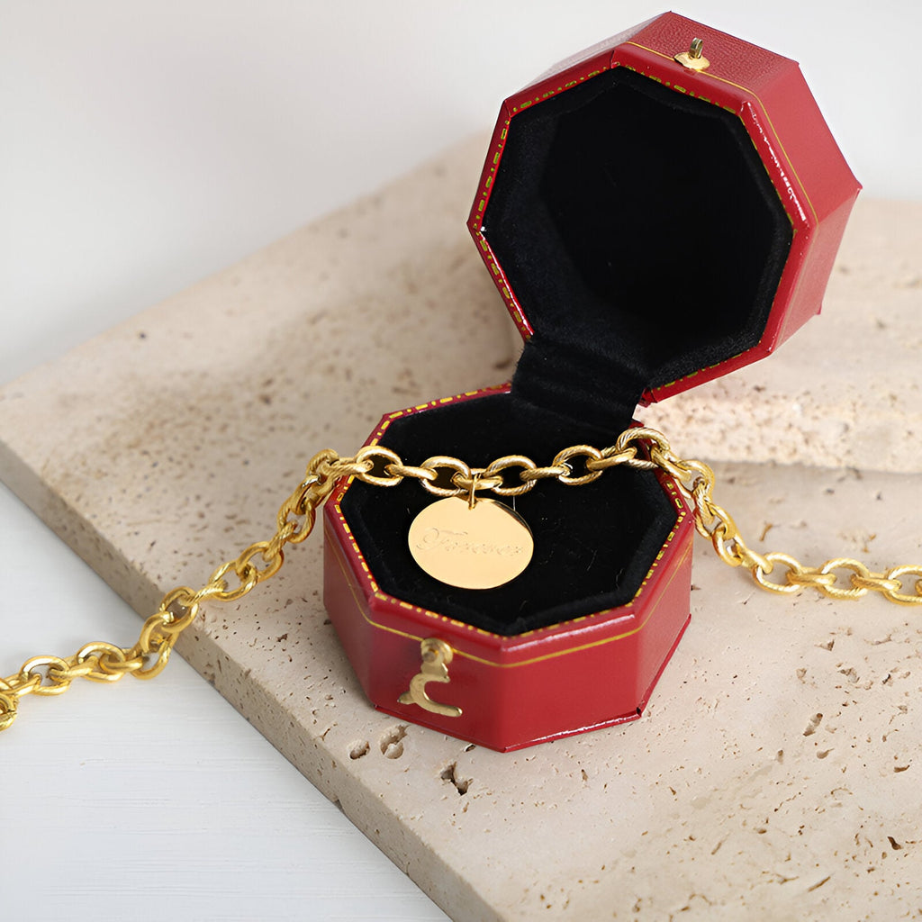 Bracelet chaîne en titane plaqué or avec médaillon rond lisse présenté dans un écrin rouge octogonal.