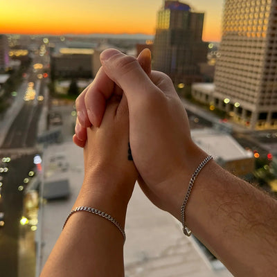 Bracelet en acier inoxydable argenté à maillons fins porté par deux poignets entrelacés au coucher du soleil.