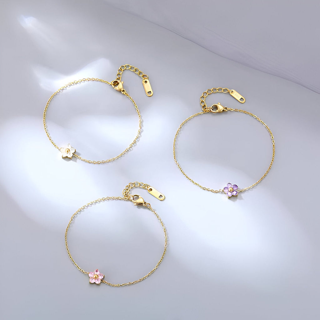 Trois bracelets en acier inoxydable doré avec pendentifs fleurs rose, violet et blanc sur chaîne fine.