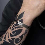 Bracelet gourmette anthracite en acier inoxydable avec plaque personnalisée et maillons larges sur poignet tatoué.