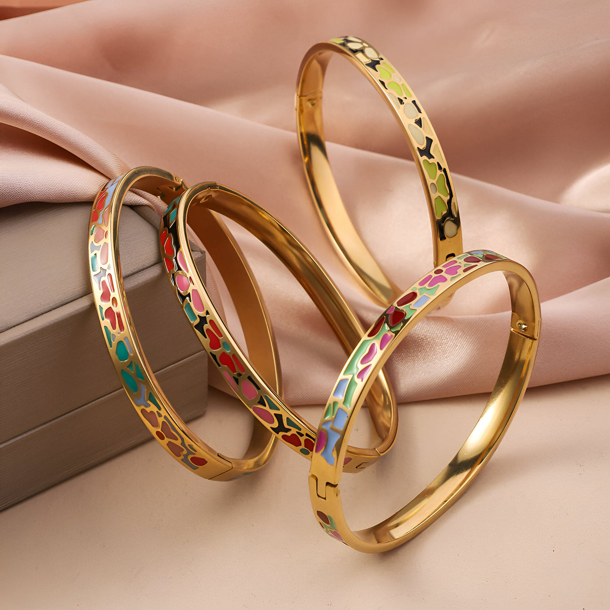Quatre bracelets joncs dorés émaillés avec motifs géométriques colorés sur fond beige satiné.