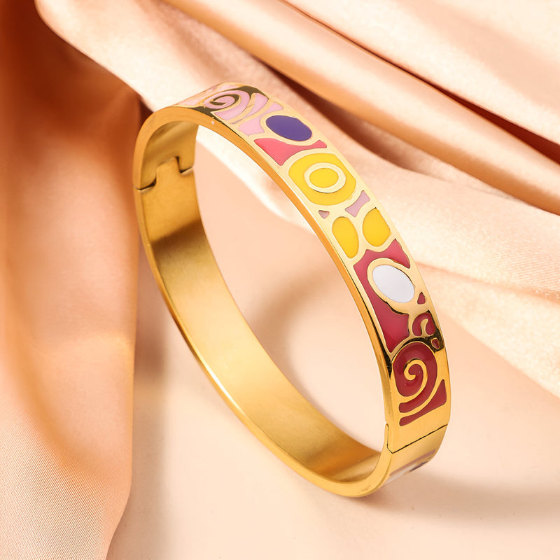 Bracelet jonc en métal doré avec motifs émaillés colorés géométriques et floraux sur fond rose pâle.