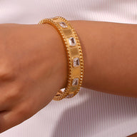 Bracelet jonc doré en acier inoxydable avec pierres carrées et détails texturés.